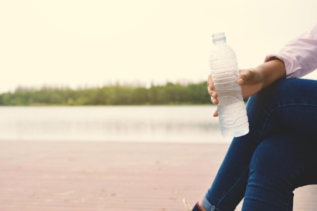 写真 明るい空の前で湖のそばに座っている女性が水のボトルを握っています