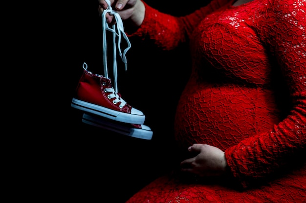 Midsection of pregnant woman holding red canvas shoes against black background