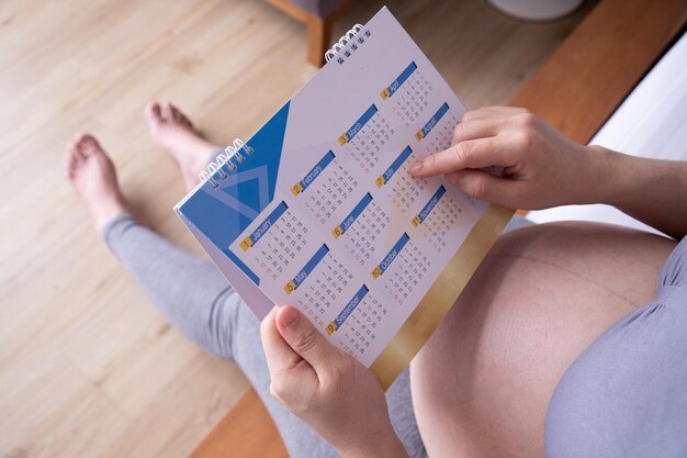 Foto sezione centrale di una donna incinta che indica il calendario mentre è seduta a letto a casa