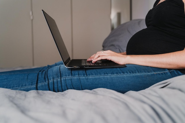 家でベッドに座っている間にラップトップを使用している妊婦のミッドセクション