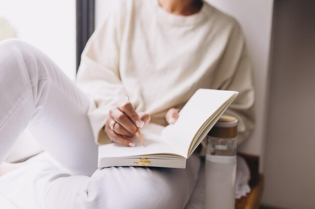 midsection of woman sitting by the window writing in a notebook