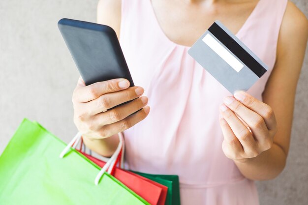 Foto sezione centrale di una donna che usa il telefono mentre tiene in mano una carta di credito e sacchetti per lo shopping mentre è in piedi contro il muro