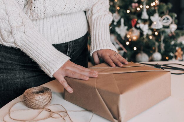 Photo midsection of woman wrapping christmas present in kraft paper in front of christmas tree at home