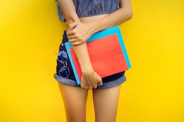 Midsection of young woman holding books while standing against yellow background