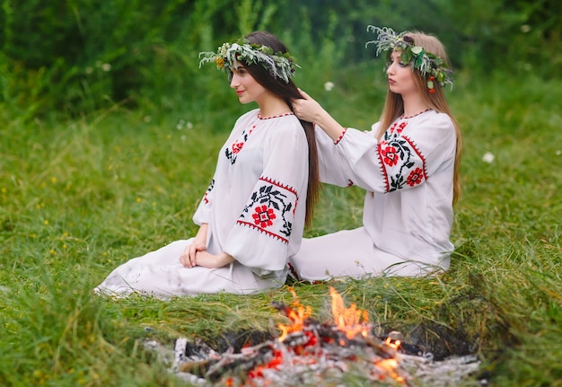 Midsummer. Two girls in the Slavic clothes weave braids in the hair near the fire.