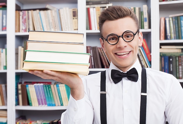 Mijn favoriete boeken. Vrolijke jonge man in overhemd en vlinderdas zittend aan de tafel in de bibliotheek en met een stapel boeken op zijn hand