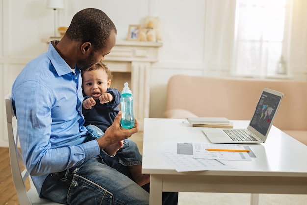 Mijn kind. Toegewijde liefdevolle jonge Afro-Amerikaanse vader die zijn zoontje en zijn fles vasthoudt terwijl hij met zijn laptop aan tafel zit