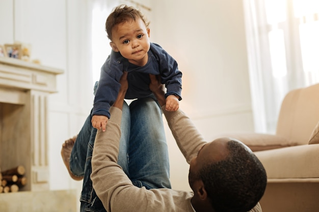 Mijn liefje. Donkerharige, bebaarde Afro-Amerikaanse man speelt met zijn vrolijke jonge zoon terwijl hij op de vloer ligt en het kind in zijn armen houdt