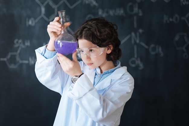 Mijn wetenschapstalent onthullen. Nieuwsgierige geconcentreerde intelligente jongen die in het laboratorium staat en geniet van scheikunde, terwijl hij deelneemt aan het wetenschapsproject en de lamp verkent