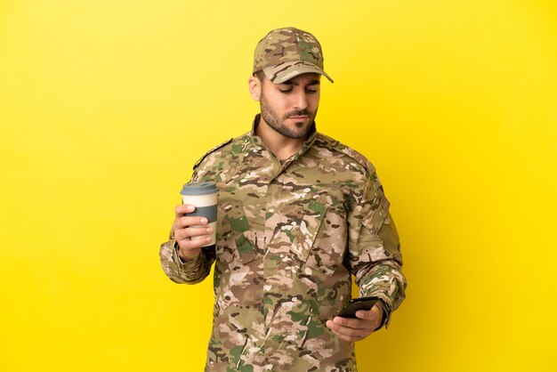 Militaire man geïsoleerd op gele achtergrond met koffie om mee te nemen en een mobiel