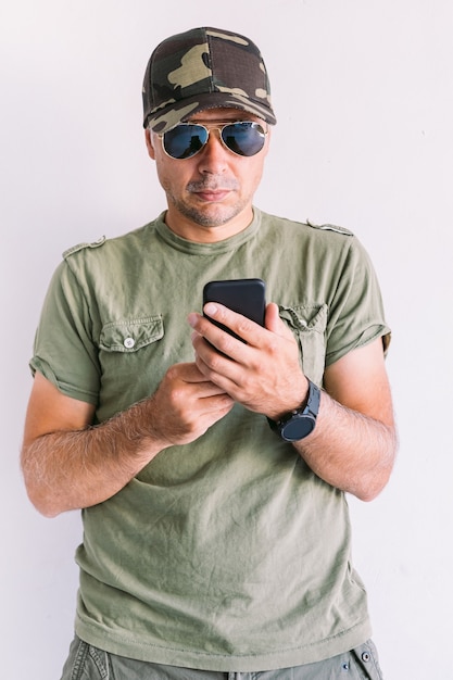 Militaire man met camouflagepet en zonnebril, kijkend naar zijn smartphone, op een witte muur