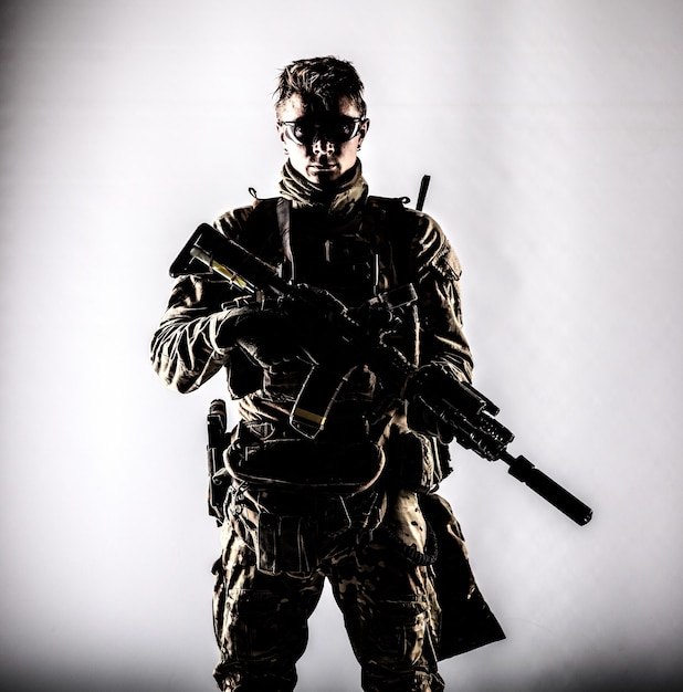 Military company mercenary, army special forces soldier, modern\
combatant in camouflage battle uniform, ballistic glasses, armed\
assault rifle with silencer low key studio portrait on white\
background