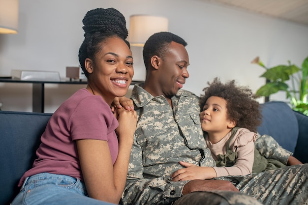 Militare, papà. giovane uomo di servizio dalla pelle scura in uniforme seduto con la moglie felice e la piccola figlia che si abbracciano sul divano in vacanza a casa