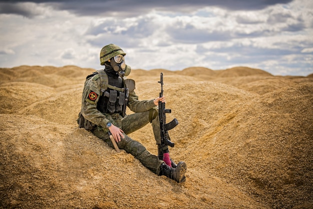 military man in a gas mask with a weapon sits saddened in the middle of the desert