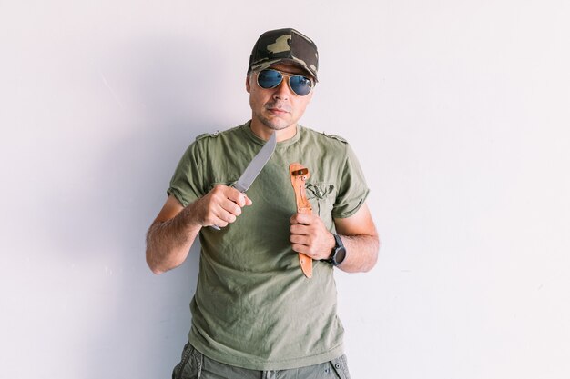 Military man wearing camouflage cap and sunglasses, with a machete, on a white wall