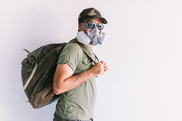 Militare che indossa berretto mimetico e occhiali da sole, con maschera di protezione e zaino militare, su un muro bianco