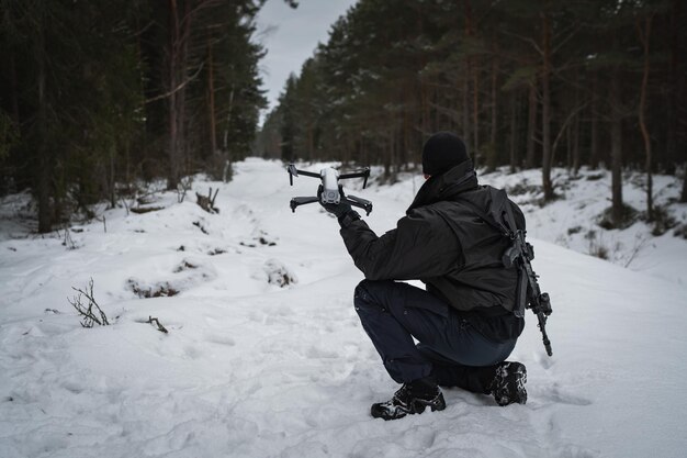 A military man with a rifle behind his back launches a drone in the forest in winter
