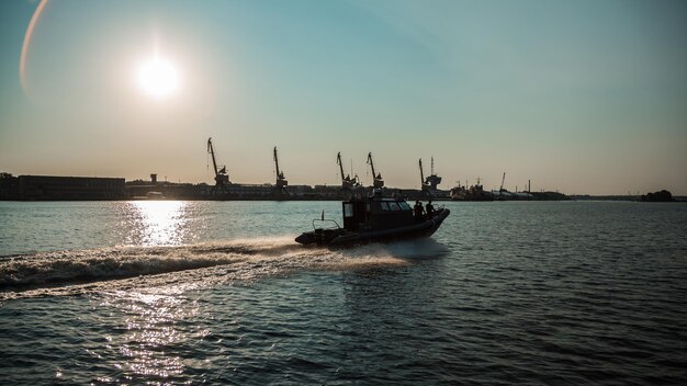 Military patrol boat on a mission in a harbor with industrial cranes and cargo ships in the background under a cloudy sky