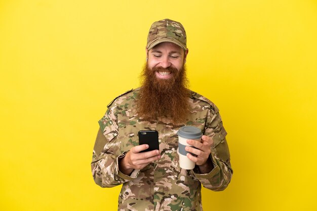 Military Redhead man over isolated on yellow background holding coffee to take away and a mobile