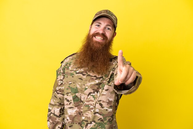 Military redhead man over isolated on yellow background showing and lifting a finger