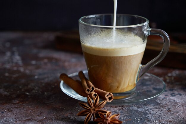 Milk foam is pouring into hot black coffee Cappuccino with sugar, cinnamon and anise on dark brown surface