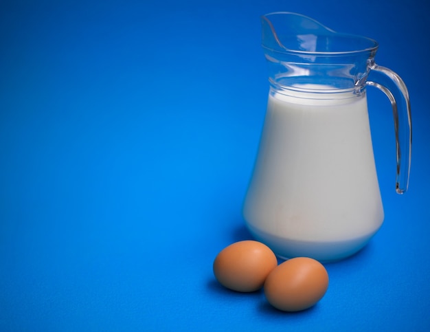 Milk in a glass jug and two eggs on a blue table