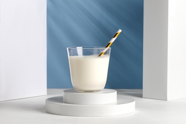 Milk in glass with a straw on white podiums. Natural milk on pedestal in sunlight. Shadow on blue wall.