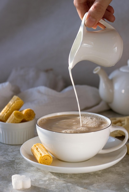 Milk is poured from the milkman into a white mug with tea on a white table among waffles