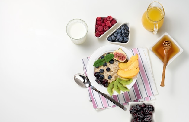 우유, 오트밀 죽과 과일을 곁들인 접시, 투명한 유리 디캔터에 갓 짜낸 주스, 흰색 테이블에 있는 그릇에 꿀. 건강한 아침 식사