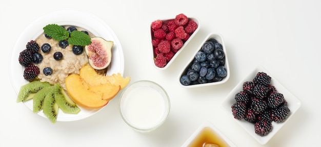 Milk, plate with oatmeal porridge and fruit, freshly squeezed juice in a transparent glass decanter, honey in a bowl on a white table. Healthy breakfast