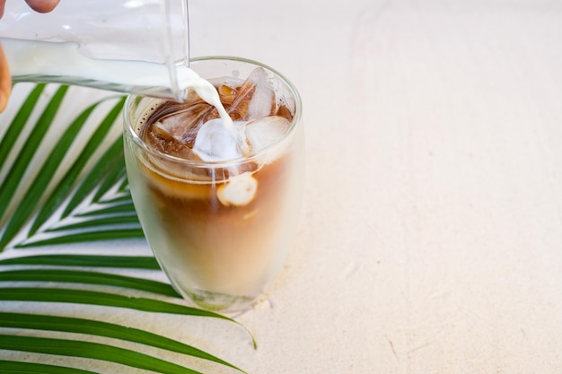 Milk Pouring Into Iced Black Coffee On The Sand Beach