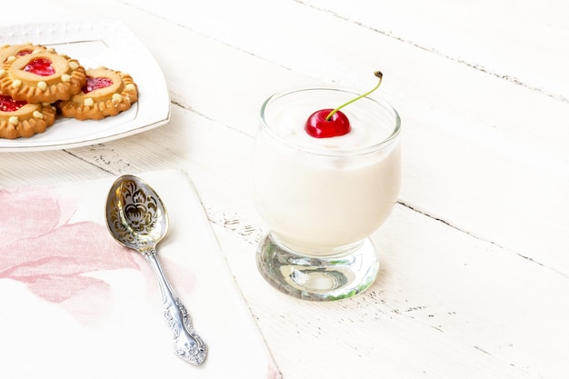 Молочный йогурт с вишней в стакане и печенье с сердечками на блюдце на белом деревянном столе крупным планом