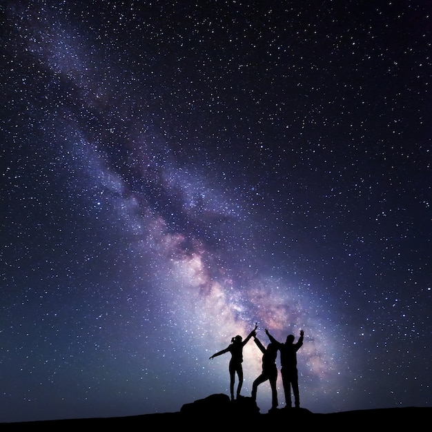 Milky Way. Night sky with stars and silhouette of a happy family on the mountain with raised-up arms.