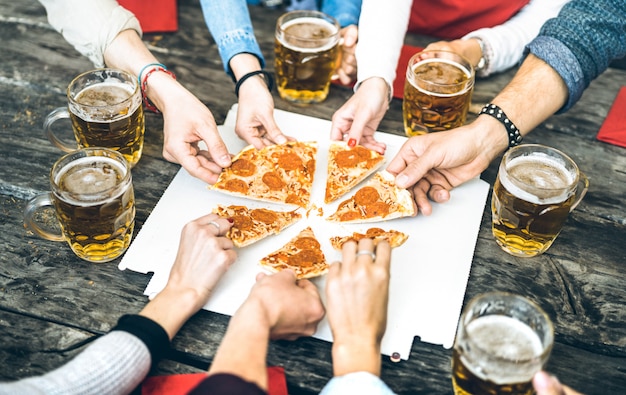 Millenial friends group drinking beer and sharing pizza slices at bar restaurant