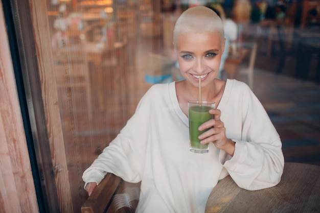 Millenial jonge vrouw blond kort haar binnen met groene smoothie