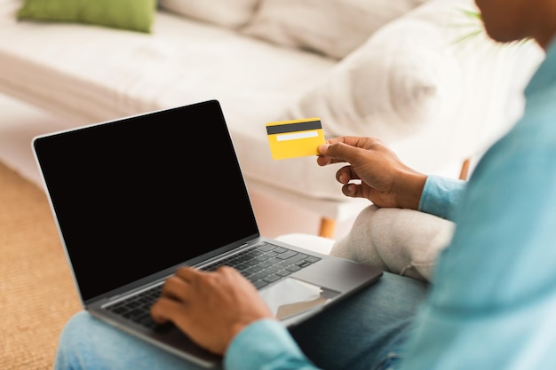 Millennial african american guy with credit card and laptop with blank screen in white living room interior