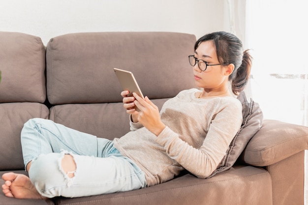 Millennial asian woman with glasses lying on the sofa using tablet to browse internet.