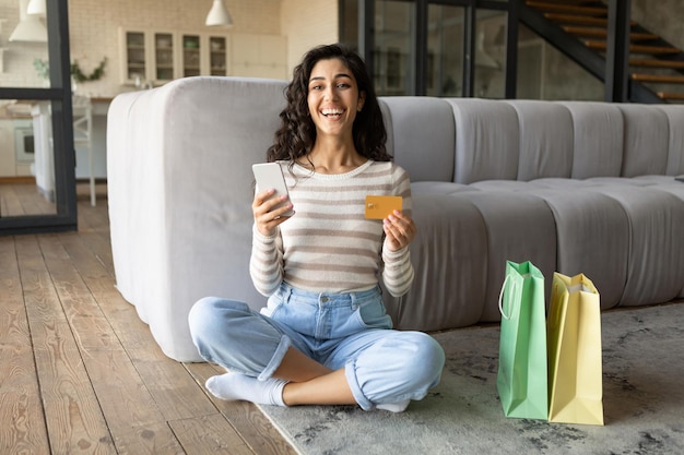 Millennial Caucasian lady purchasing goods over internet using smartphone and credit card at home free space