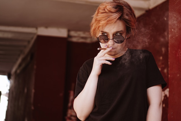 Millennial cool pretty girl with short red hair and mirror sunglasses smoking cigarette in the old city with red walls