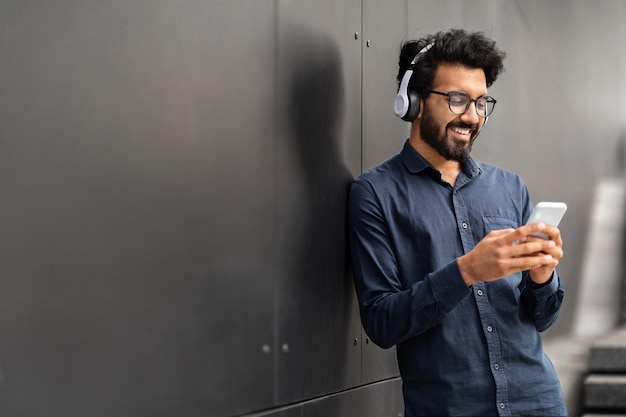 Millennial eastern man standing by black wall using phone headphones