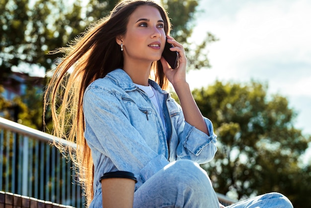 La ragazza millenaria in vestiti di jeans si siede su una panchina nel parco, comunica con amici, colleghi, genitori al telefono.