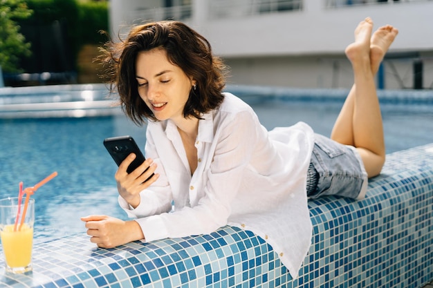 A millennial girl lies with a phone on the edge of the swimming\
pool in a man\'s shirt and communicates on the internet in social\
networks with friends online technologies and communications