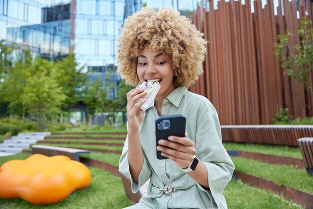 Millennial girl with curly blonde hair uses cellphone chats
with friends online types text message bites delicious sweet snack
wears dress smartwatch poses outdoors in urban park reads good
news