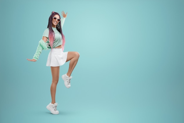 Millennial girl with pink braids hairstyle in turquoise clothes and sunglasses posing on a blue background. Concept for modern style, inclusiveness, individuality, generation Z, copy space.