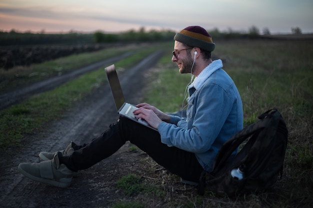 Millennial hipster man working on laptop with earphones in field at sunset