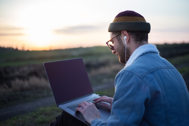 Millennial hipster man working on laptop with earphones in field at sunset