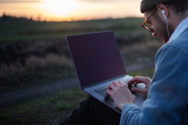 Millennial hipster man working on laptop with earphones in field at sunset