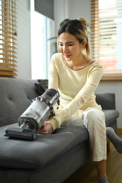 Millennial housewife cleaning sofa with cordless vacuum cleaner to removes germs and dirt Housework concept