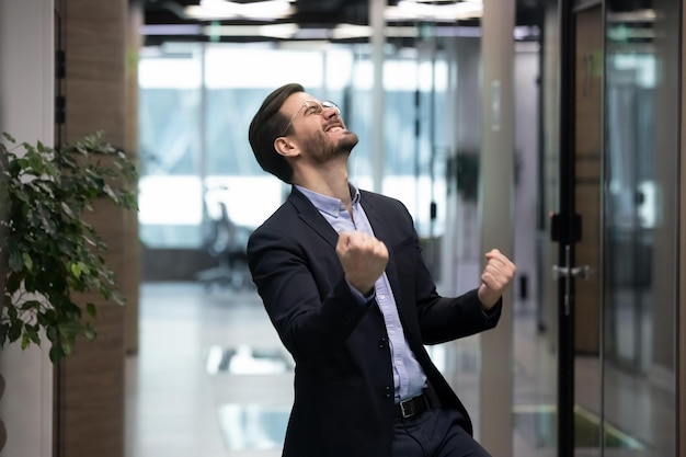 Millennial office worker in suit standing in hallway clenched fists feel overjoyed celebrate career advancement got business opportunity salary or sales growth successful winner businessman concept