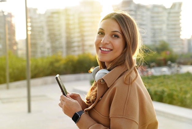 Millennial. portrait female. teens looking at camera. mobile\
app. headset phone. smartwatch. sunlights rays. sunset. winter\
clothes. urban background.